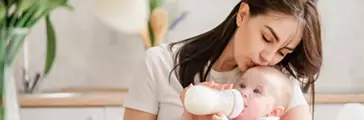 Young mother kiss baby during drinking milk. Nursing a baby. Feeding newborn with formula in a bottle.