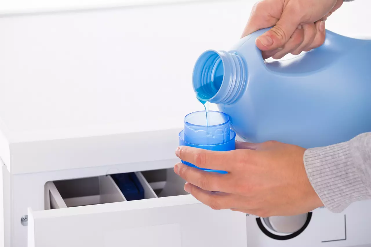 Cropped image of young woman's hand pouring detergent in lid with washing machine in background